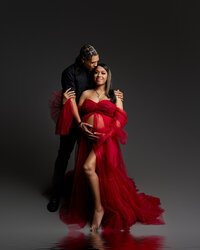 Expecting couple posing together during a dramatic studio maternity photoshoot with mother wearing a flowing red tulle maternity gown while father embraces her and holds her baby bump, photographed by Kaitlyn Dawn Photography.