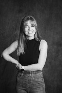 A black and white picture of a blonde woman with straight hair smiling in front of a dark gray backdrop. She wears jeans and a black tank top.