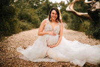 A pregnant woman in a princess style gown smiles with her arms above and below her bump. She sits on a mulched path with bushes on either side. A curved tree shows in the background.