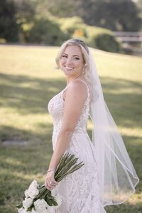 A St. Augustine bride smiles softly over her shoulder while holding her bouquet stems, captured in natural light. Her fitted lace gown features delicate detailing, and her long veil flows gently behind her in the afternoon breeze. With soft waves tucked behind one ear and minimal jewelry, her look is both refined and effortless. Photographed by Phavy Photography in a true-to-color, documentary style that highlights timeless elegance and authentic bridal beauty.