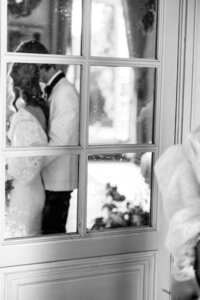 Black and white photo of a bride and groom sharing an intimate moment, seen through a window with soft reflections, creating a romantic and timeless wedding portrait.