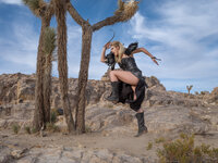 Model in dark clothing and boots posed on desert rock among Joshua trees under a bright blue sky.