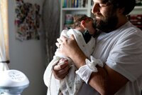 Professional photography by Nature's Child Photography: Mandarin lifestyle newborn session capturing a quiet in-home moment as a father holds his baby girl, her soft headband looped around his arm while she rests peacefully against him.
