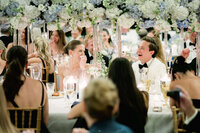 Couple laughing together at their reception table beneath a canopy of white and blue florals as guests smile around them.