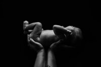 A black and white picture of a newborn in his parent's hands. he lays against a black backdrop and appears to be held up to a light.