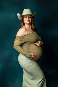 Expecting mother wearing a green off shoulder maternity gown and cowboy hat during a dramatic studio maternity portrait session by Kaitlyn Dawn Photography.