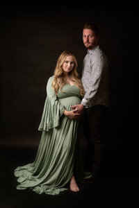 Expectant couple embracing, wife in a sage green gown, during a Cleveland maternity photography session in a dark studio
