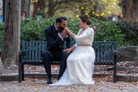 a newlywed couple sitting on a bench as one kisses the other's hand