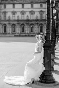 Black and white photo of a bride in a fitted lace gown leaning against an ornate lamppost in an open Paris square, with classic architecture in the background.