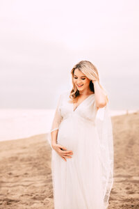 A blonde pregnant woman stands on a beach. She smiles brightly towards her bump. She holds one hand underneath it while fluffing her hair with the other. The sky and beach are bright on an overcast day.
