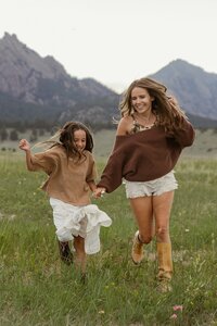 Mother and daughter frolic in a grassy meadow at golden hour with Flatirons mountain backdrop during a family photography session near Boulder, Colorado.