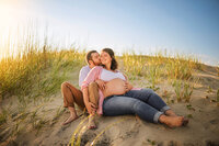 Expecting couple embracing on sand dunes at sunset during maternity photography session near the North Carolina coast