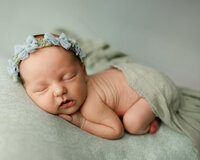 Sleeping newborn baby girl wearing a floral headband and posed on a soft green backdrop during a studio session with newborn photographer Kaitlyn Dawn Photography.