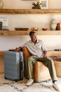 Model in a linen shirt and olive trousers seated beside grey MUJI luggage