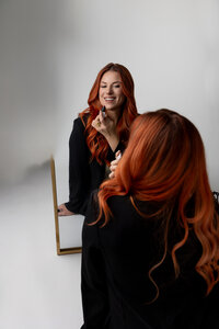 Red-haired woman smiling while applying lipstick and looking at her reflection in a mirror during a studio branding session. Photograph by Yucaipa branding photographer Kaitlyn Dawn Photography.