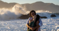 man holding woman from behind while kissing her at sunset with the large waves crashing against the rocks in the background
