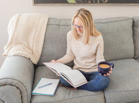 A relaxed business owner sits cross-legged on a couch, reading a book with a blue mug in hand and a notebook nearby—ideal for coaches and entrepreneurs creating content from home.