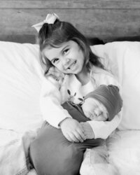 Black and white portrait of a smiling big sister holding her sleeping newborn sibling during an in-home lifestyle newborn session.