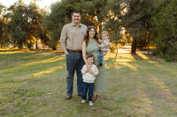 Parents standing with their two children in a grassy park at sunset, soft backlighting and relaxed smiles captured by Kaitlyn Dawn Photography, professional family photographer.