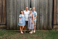 Family of four standing together in front of a wooden barn during a family portrait session in New Bern