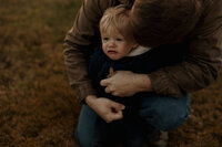 Maine family photographer capturing a candid moment of a father helping his young son zip up his jacket while hugging him close, with the boy looking slightly sad in a tender, authentic lifestyle portrait