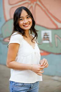 Smiling high school senior girl in a white blouse standing in front of a colorful mural, outdoor senior portrait by Kaitlyn Dawn Photography.