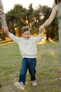 Child lifted joyfully by his parents’ hands during golden hour in a sunlit field, candid outdoor family portrait captured by Kaitlyn Dawn Photography.