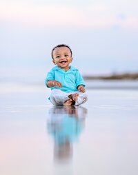 Professional photography by Nature's Child Photography: Laughing baby boy in a teal polo sitting barefoot on wet sand as his reflection mirrors him in the shallow water during a beach child photography session in Palm Coast.