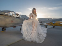 Woman in a sheer embellished gown standing before a vintage twin-engine aircraft on a runway at sunset.