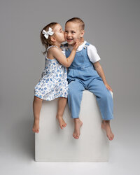 Two siblings sitting on a white studio cube, little girl kissing her brother in coordinated blue outfits, photographed by Kaitlyn Dawn Photography, children’s and sibling portrait photographer.