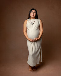Expecting mom in a neutral beige dress standing against a warm backdrop, showcasing her pregnancy during an Inland Empire maternity session with Kaitlyn Dawn Photography.