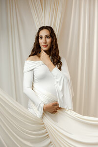 Expecting mom in a white maternity dress with elegant draping, gently holding her belly during a maternity session in Yucaipa by Kaitlyn Dawn Photography.