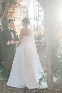 The bride and groom stand beneath the floral-covered chuppah, bathed in warm, golden light
