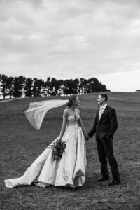Black and white image of bride and groom looking at each other holding hands. Hill lined with trees in the background.