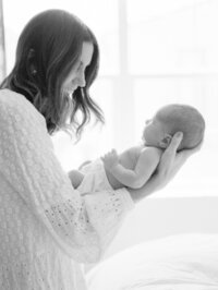 Black and white portrait of a mother holding and admiring her newborn baby beside soft window light during an in-home session.