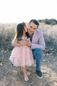 Young daughter from the Parker family kissing her father on the cheek while he smiles, captured in a warm desert setting in Las Vegas during a natural light family photography session.