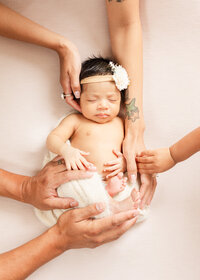 Lifestyle newborn photo—baby swaddled with parents’ and sibling’s hands gently surrounding her