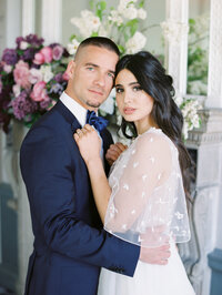 bride and groom looking at the camera with floral installation on fireplace and detailed bridal gown visible