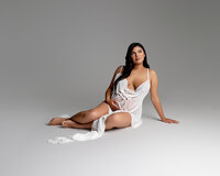Pregnant woman sitting on the studio floor wearing a flowing white maternity dress during a studio maternity portrait session by Kaitlyn Dawn Photography.