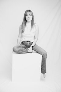 A black and white picture of a blonde woman with straight hair sitting atop a white box in front of a white backdrop. She wears a white shirt and jeans. One leg rests off the side of the box as she tucks the other beneath her, resting a hand on her foot.