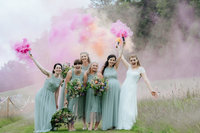 bride and her bridesmaids stand in a feild at Hadsham Farm Wedding venue and wave colourful smoke bombs.  the smoke bombs make pink, white and orange smoke plumes whilst the girls smile towards the camera