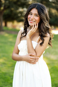 High school senior girl smiling in a sunlit park wearing a white dress, close-up senior portrait by Kaitlyn Dawn Photography.