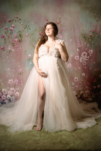 A pregnant woman in a tulle dress smiles up and out of the frame. She has one arm bent against her chest with her hand in her hair while the other arm drapes around her bump, holding it from beneath. She stands in front a multi-colored floral backdrop. One of her legs is forward, peaking out of the dress.
