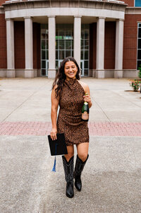 Girl wearing black cowboy boots and leopard print dress carrying a bottle of champagne and graduation cap