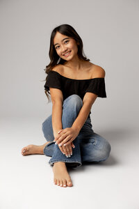 High school senior girl in a black off-shoulder top and jeans sitting on the floor in Yucaipa studio portrait session by Kaitlyn Dawn Photography