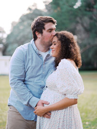 Husband is kissing his pregnant wife on her temple while she smiles, his hands on her belly