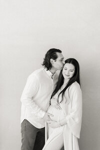 Black and white in studio maternity portrait of an expecting Dallas mother being held and kissed by her spouse.