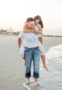 In Palm Coast, an engaged couple laughs during a beach as she wraps her arms around his shoulders and he carries her barefoot along the water during their engagement announcement. - A portrait experience by Nature's Child Photography.