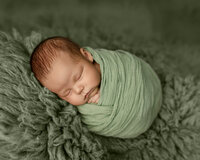 Sleeping newborn baby boy wrapped in a soft green swaddle and posed on a sage green backdrop during a studio newborn photography session by Yucaipa newborn photographer Kaitlyn Dawn Photography.