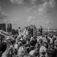 Cérémonie de mariage au Belvédère du Vieux-Port de Montréal, couple sous arche florale avec invités et skyline en arrière-plan, portrait en noir et blanc